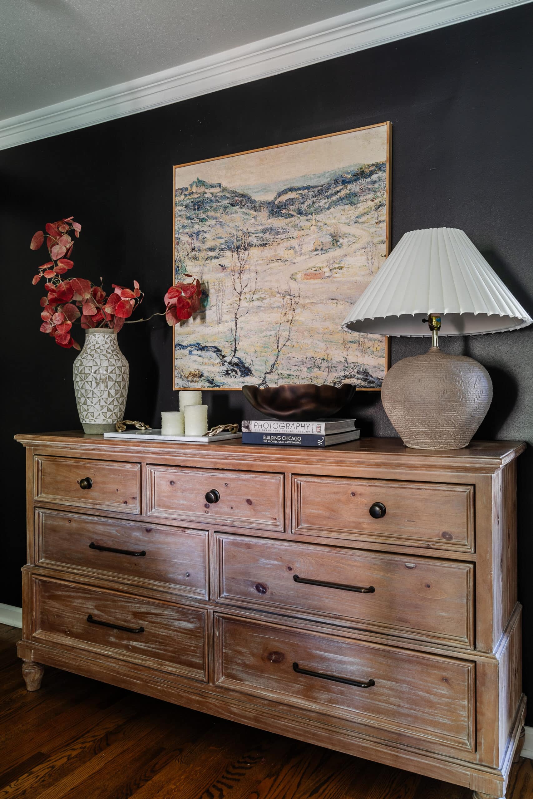 wood dresser with large wall art, vase of fall branches, and table lamp