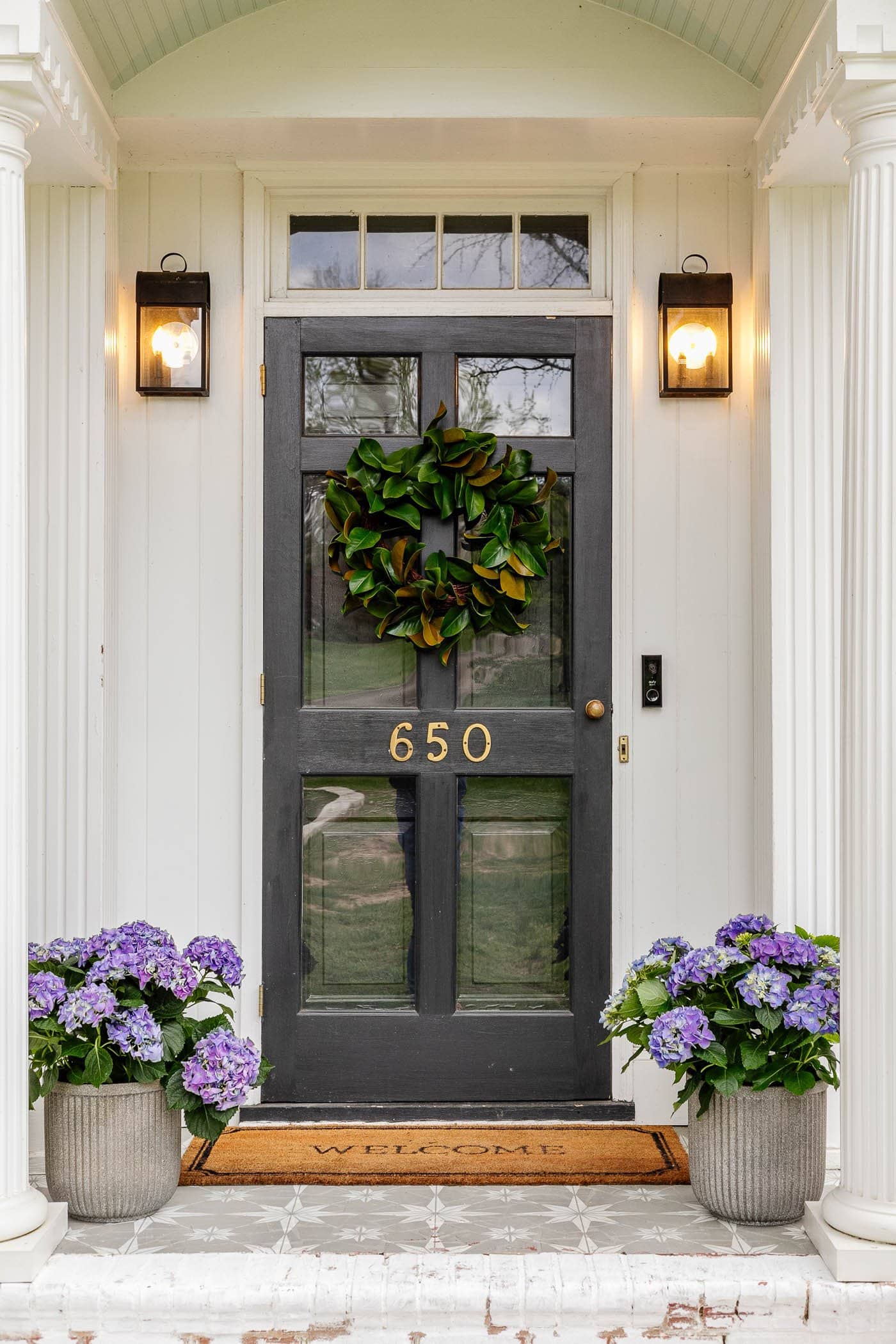 black front door with white columns on a small front porch with magnolia wreath and purple hydrangeas and an extra wide welcome mat