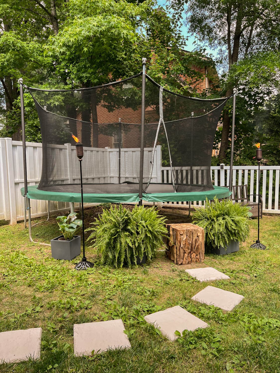 stepping stones leading to outdoor trampoline with tree stump trampoline ladder, ferns, and tiki torches