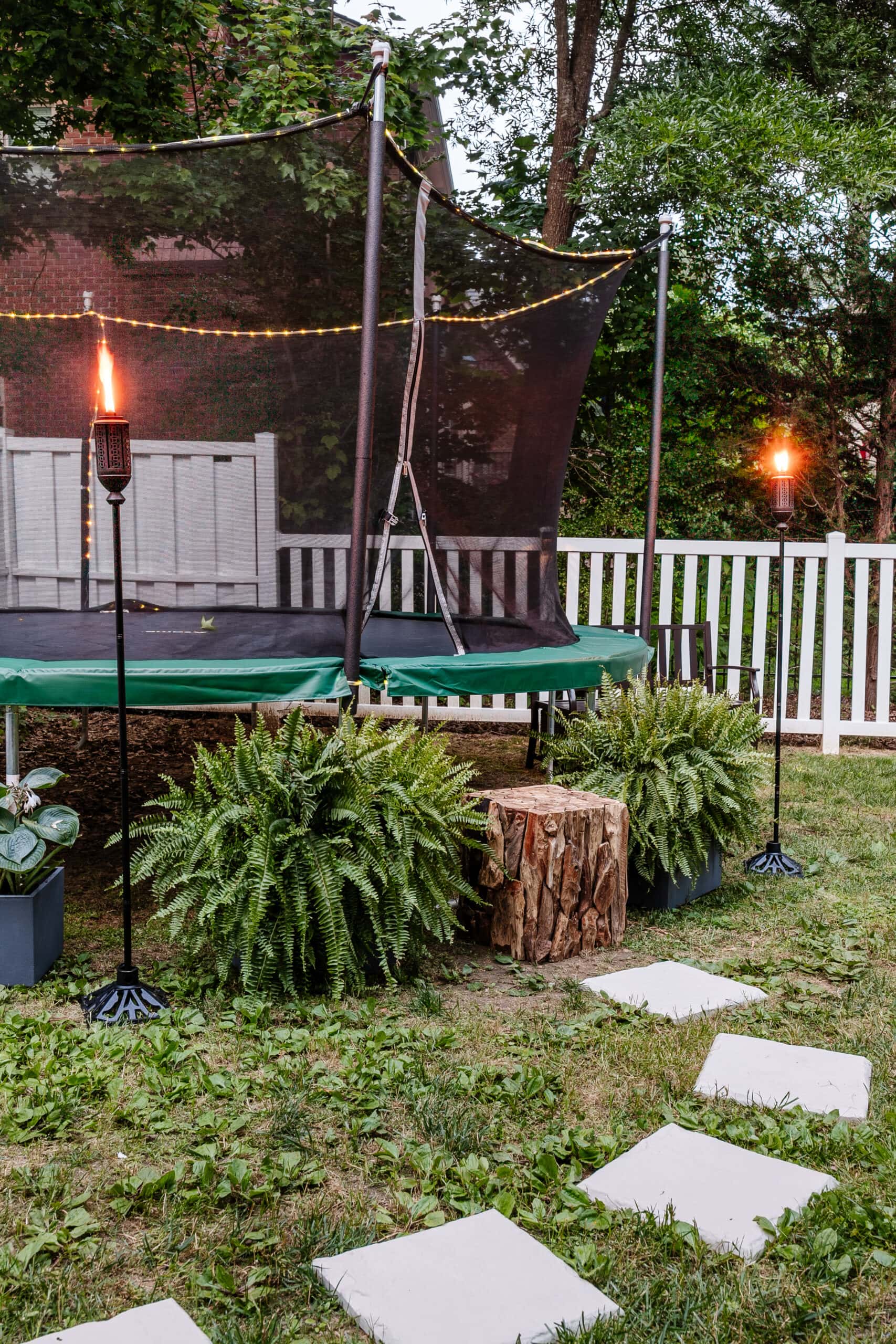 stepping stones leading to outdoor trampoline with tree stump trampoline ladder, ferns, and tiki torches