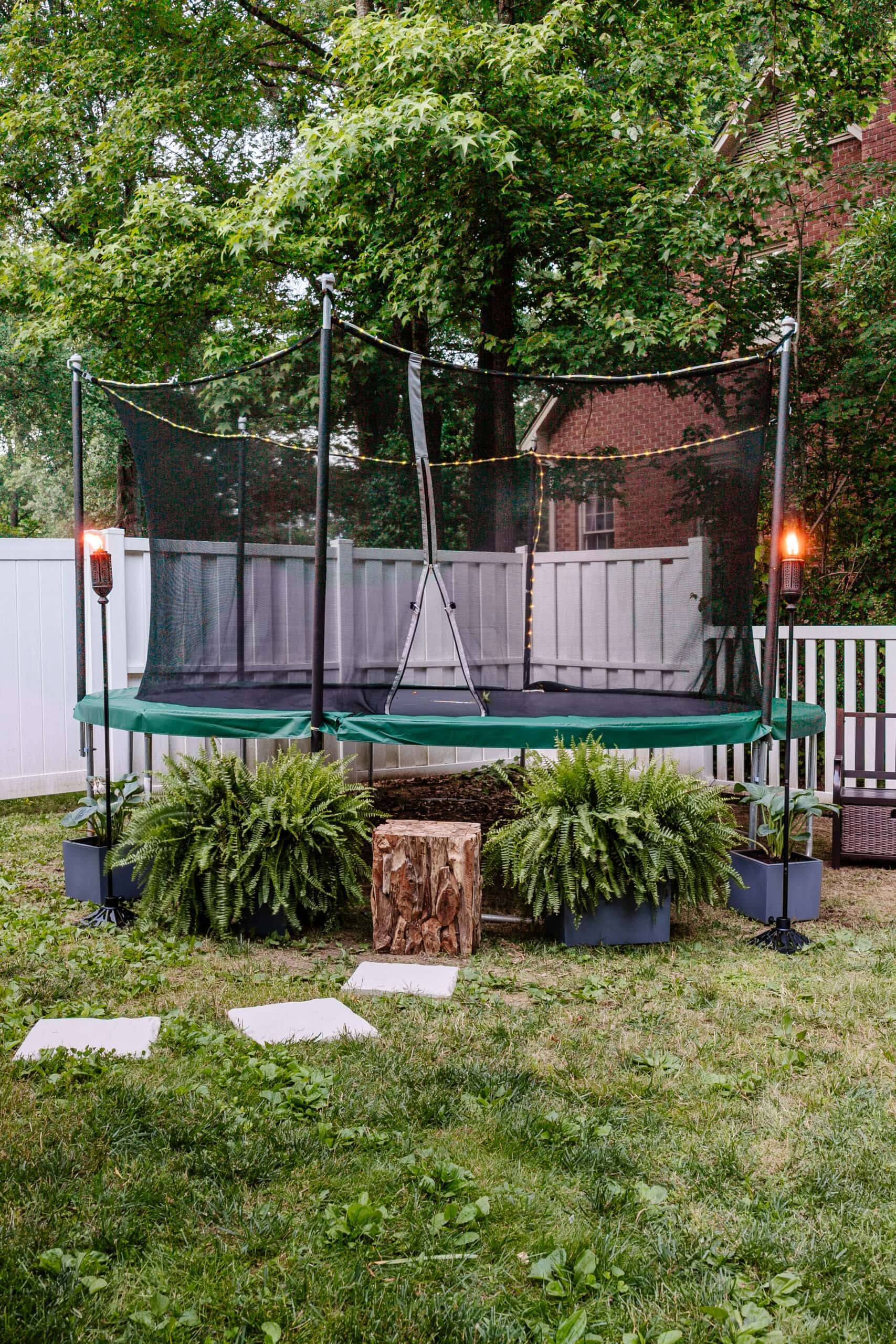 stepping stones leading to outdoor trampoline with tree stump trampoline ladder, ferns, and tiki torches