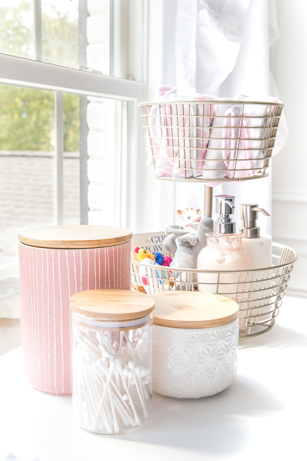 Nursery Organization | Use a tiered fruit basket from the kitchenware aisle to store baby washcloths, grabby toys, Wubbanubs, lotion, and hand sanitizer and lidded kitchen canisters to store Q-tips, pacifiers, and hair bows.
